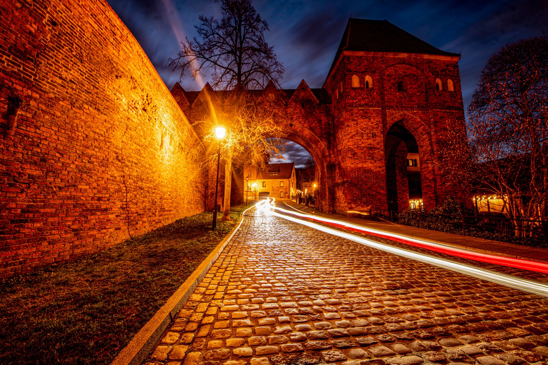 Stunning long exposure photograph capturing the historic gate in Toruń, Poland at night with vibrant light trails.