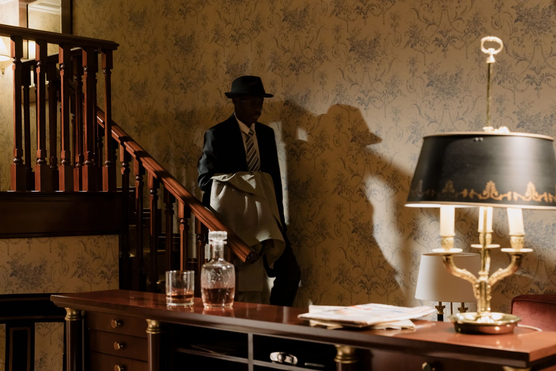 A shadowy detective in vintage attire descends wooden stairs, evoking a classic noir mood.