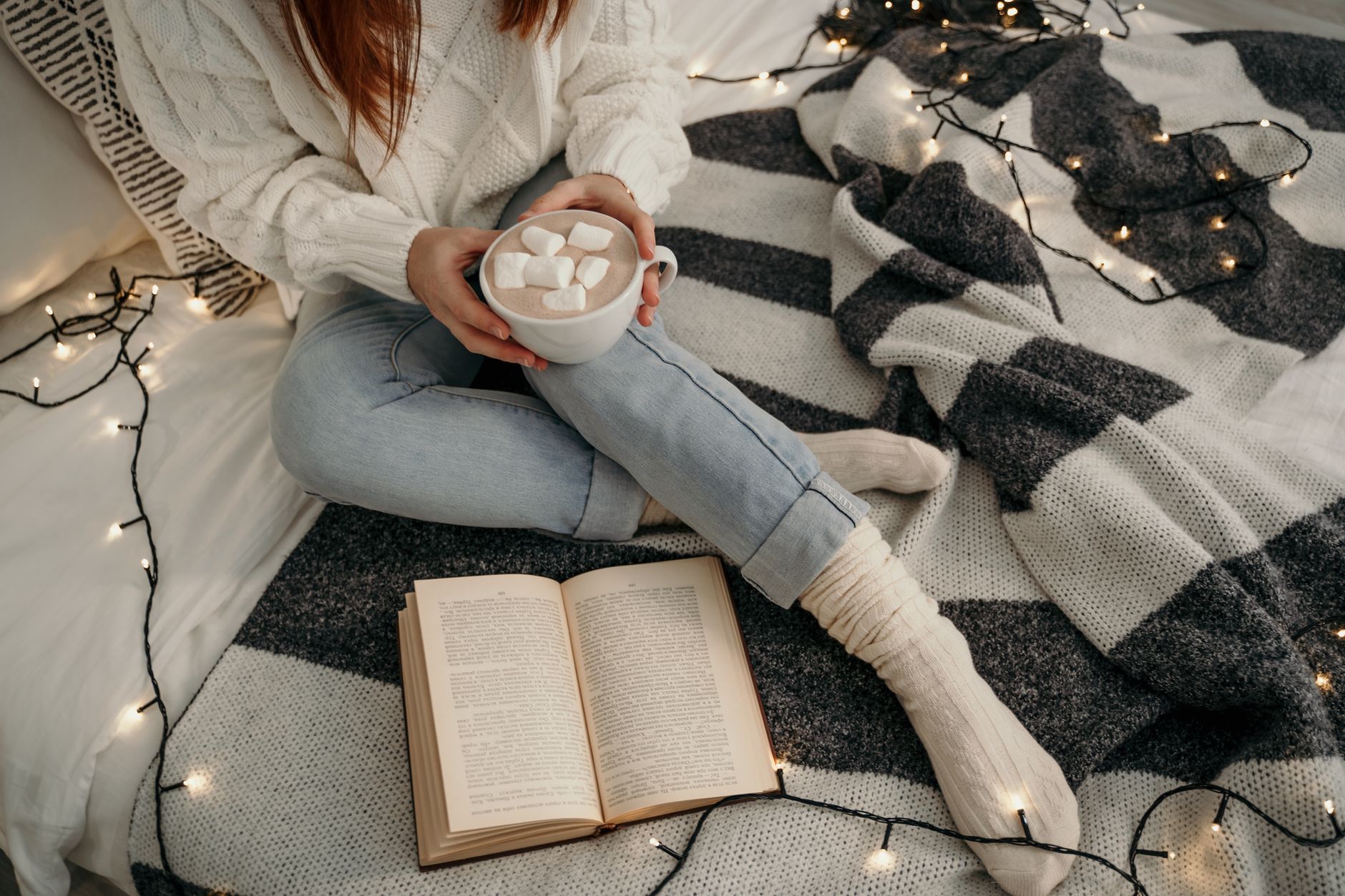 A woman enjoys marshmallow hot chocolate, sitting on a bed with fairy lights and a book, exuding cozy winter vibes.
