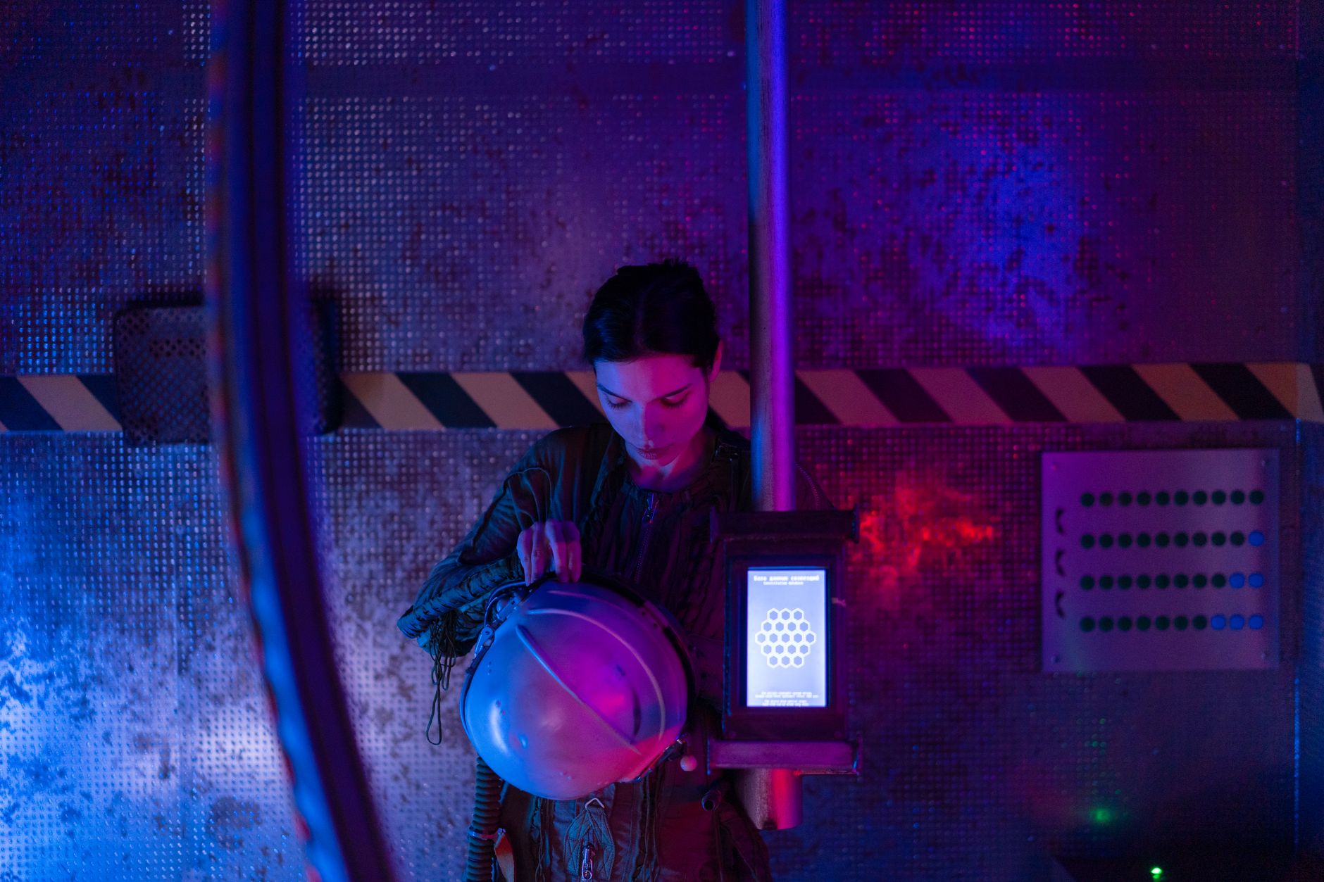 A female astronaut in a spacesuit examines her helmet in a dimly lit spaceship interior.