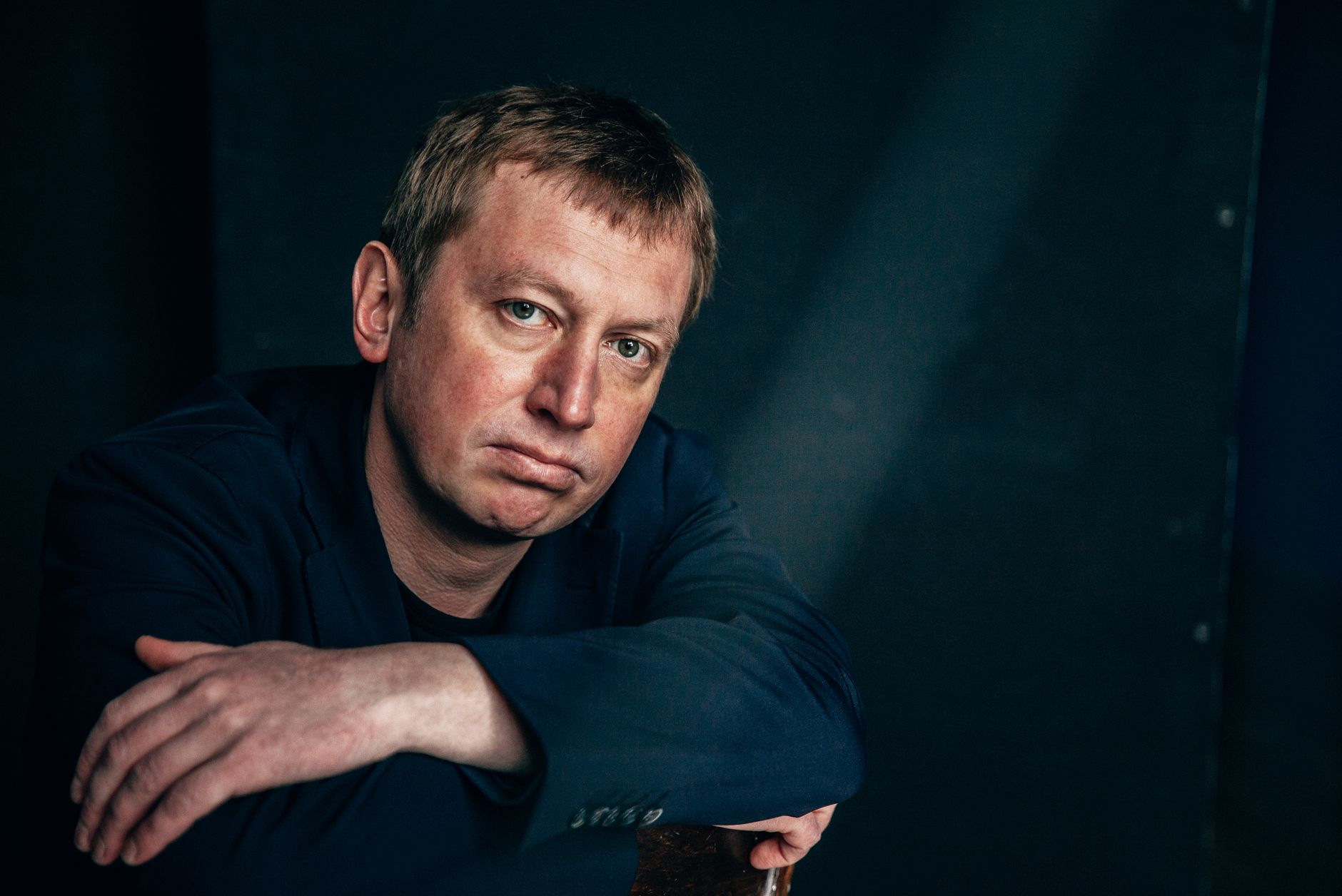 Close-up portrait of a man in a black coat, sitting against a dark wall.