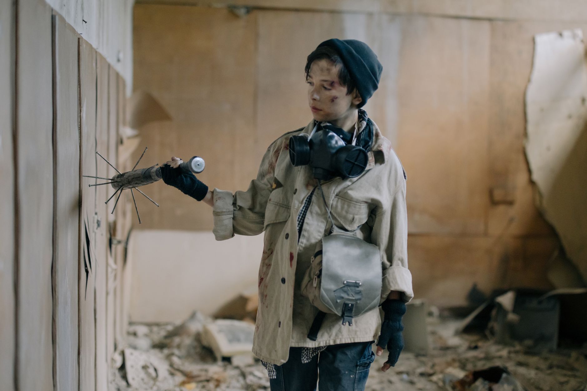 A young survivor in a gas mask with a nail bat in an abandoned building.