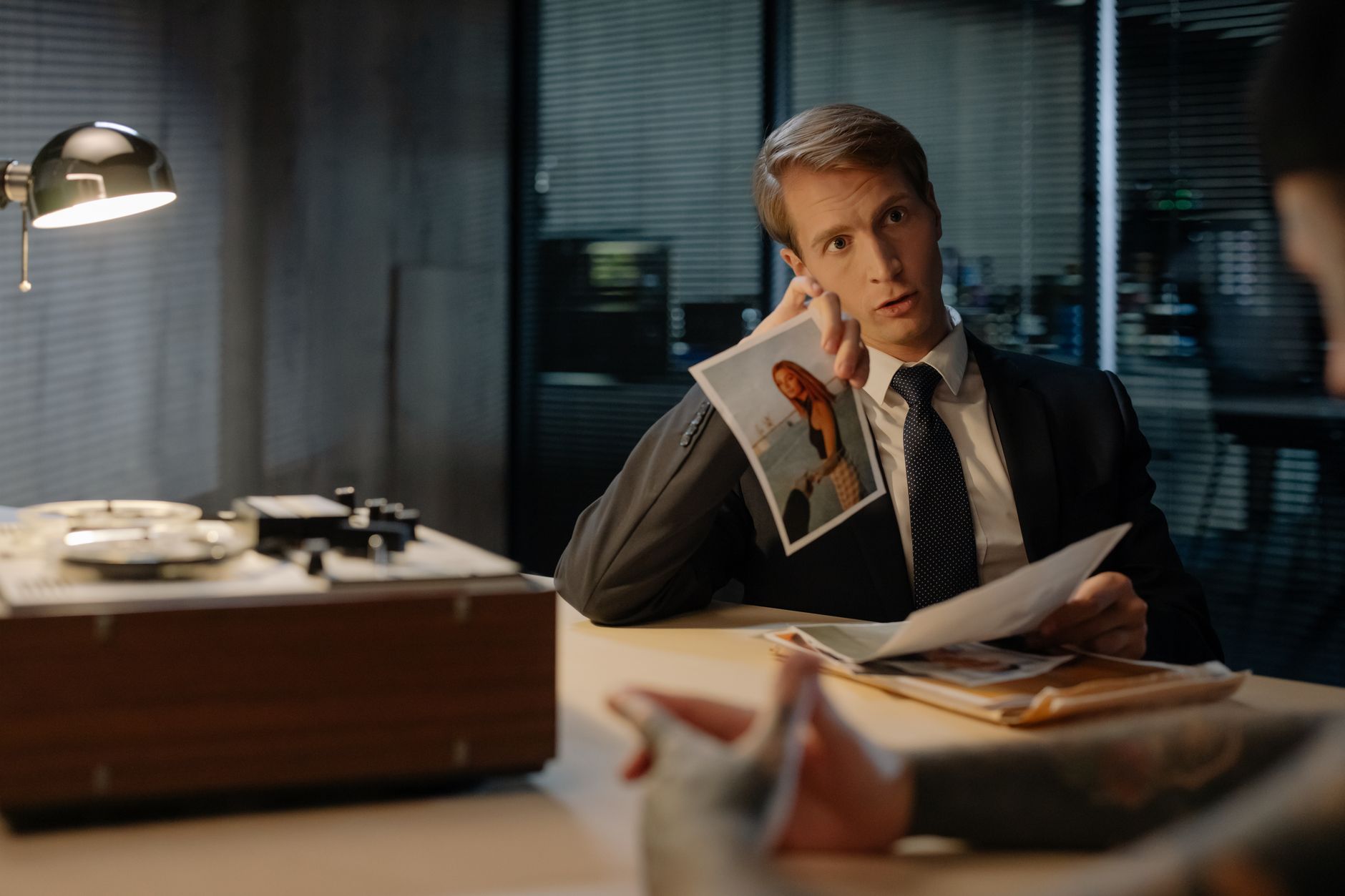 Male detective questioning suspect with evidence photos in dimly lit interrogation room.