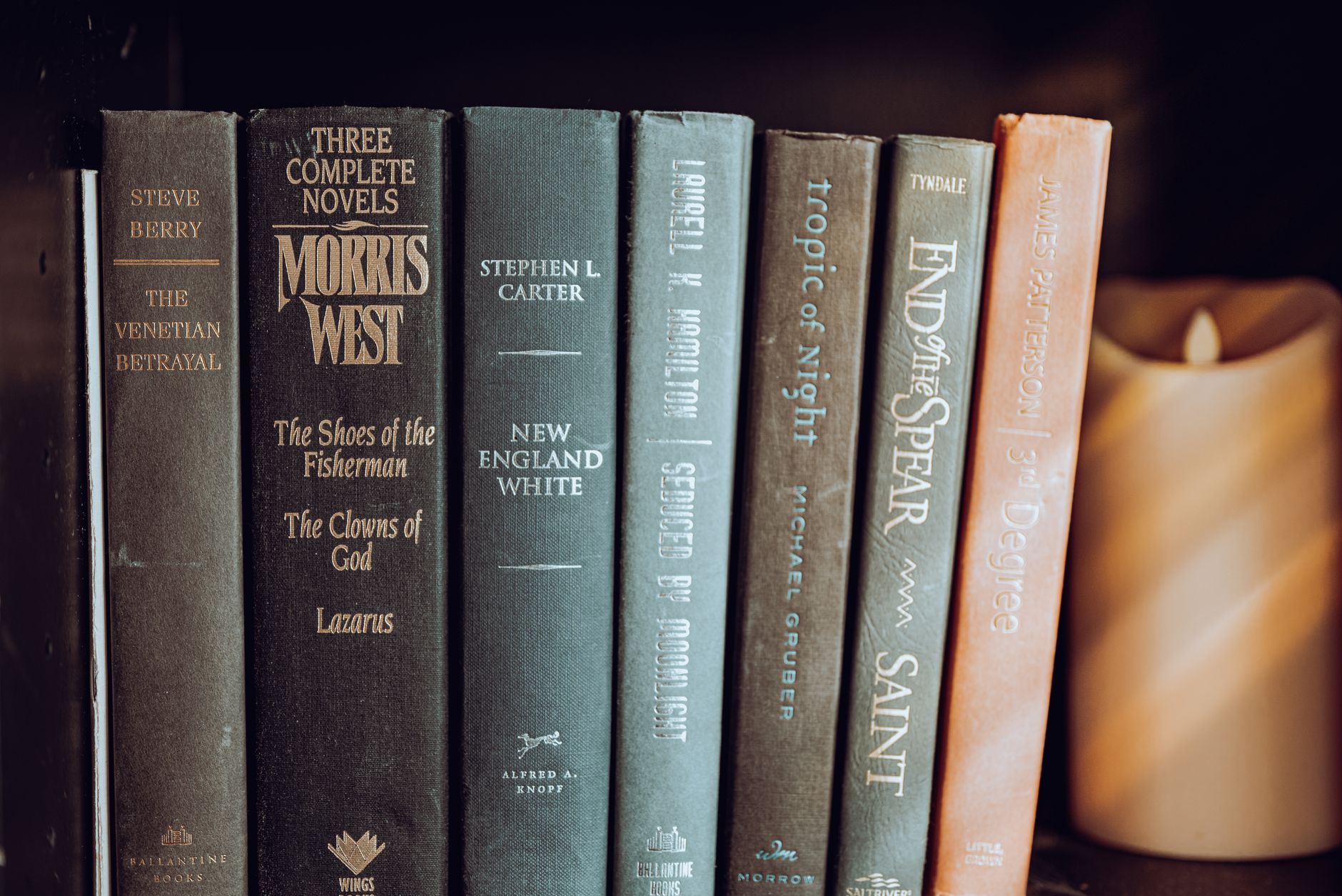 A close-up view of classic novels neatly arranged on a bookshelf with a warm-toned candle.