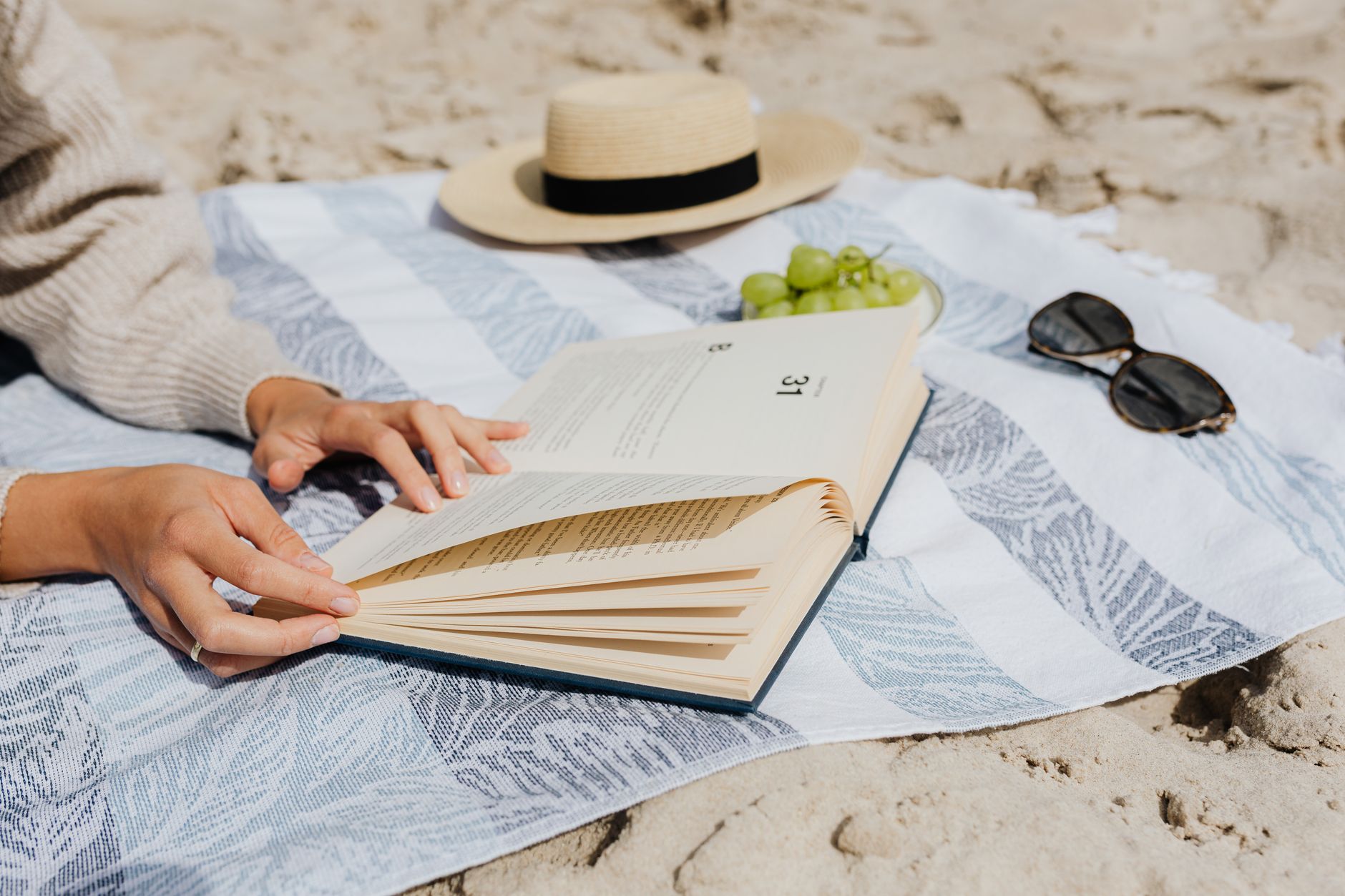 Cozy beach scene with book, grapes, sun hat, and sunglasses on a blanket.