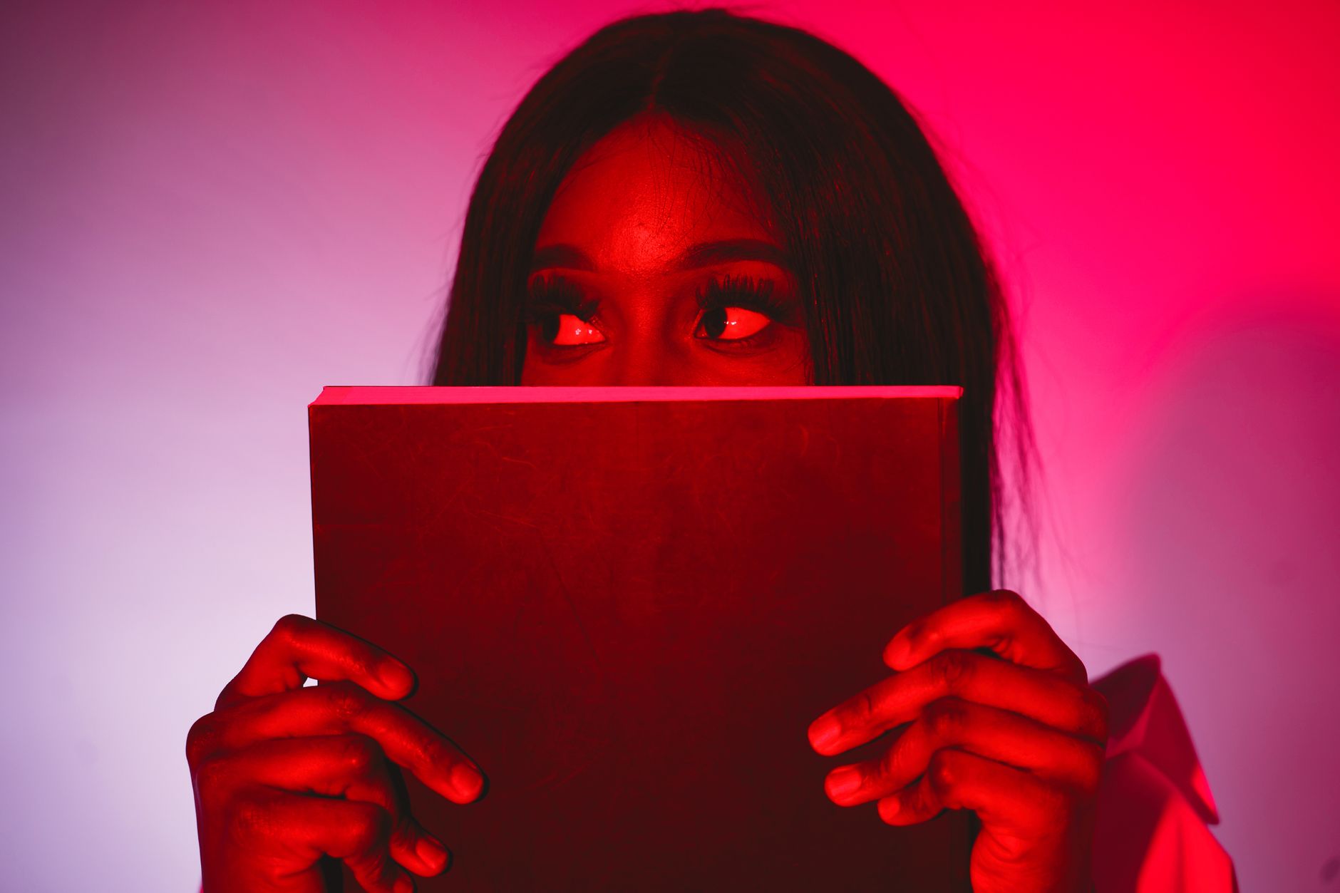 A mysterious woman with false eyelashes peeking over a book under red lighting.