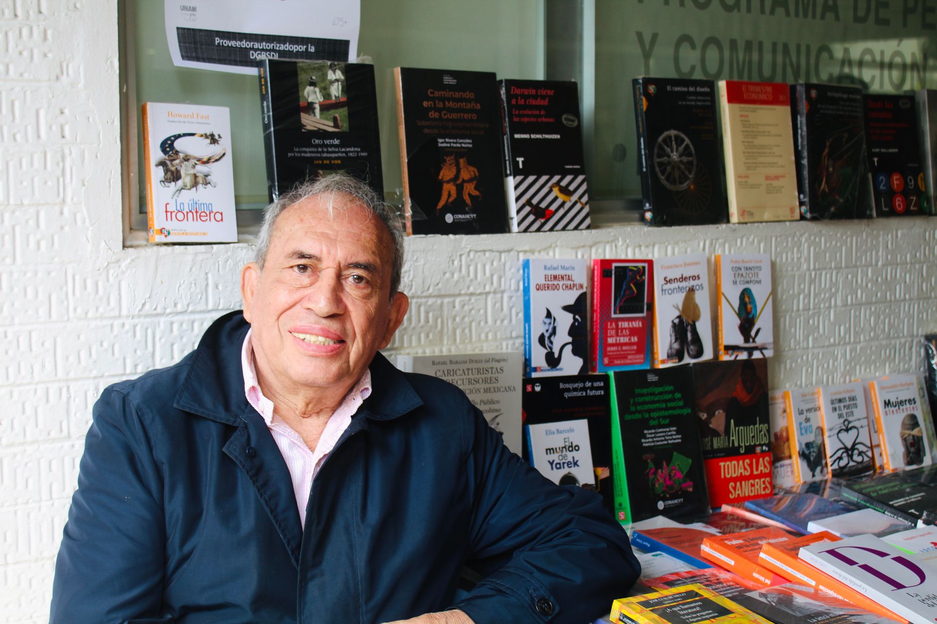 Smiling man sits by a book stall featuring a variety of published titles, showcasing his collection.