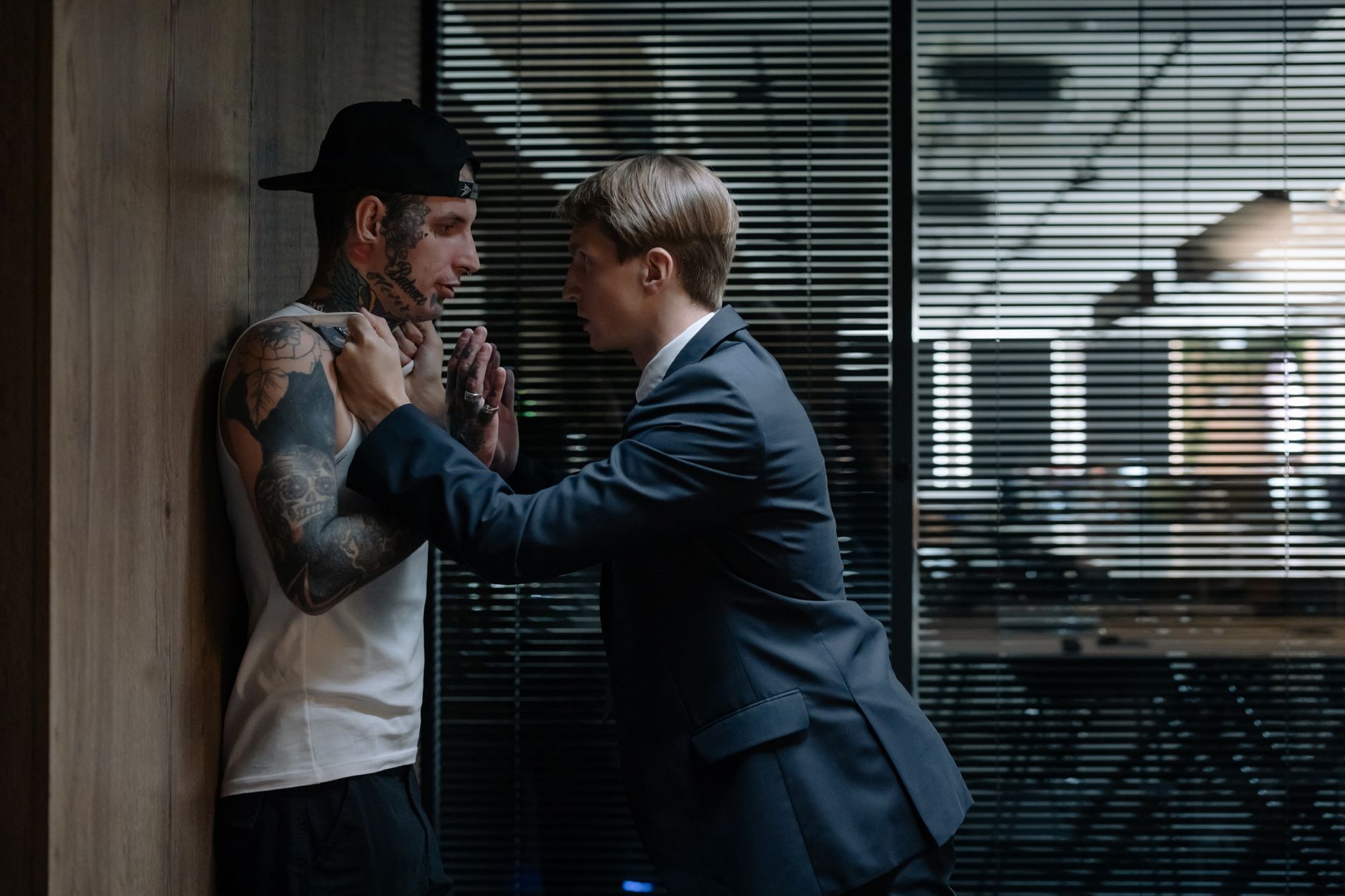 Business-suited detective confronts tattooed suspect in tense indoor setting, emphasizing investigation.