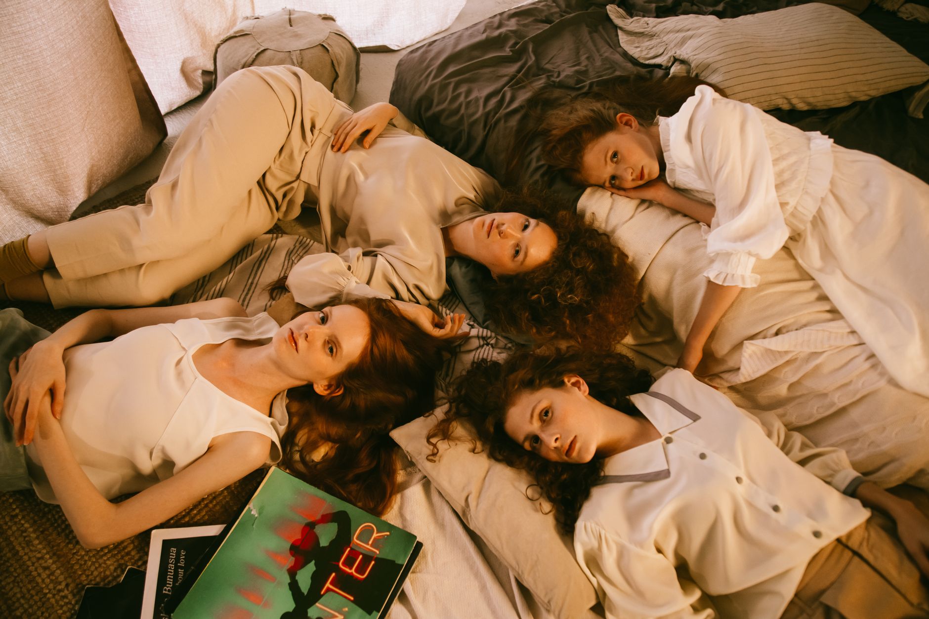 Four women enjoying a relaxed sleepover, lying on a bed with pillows and books.