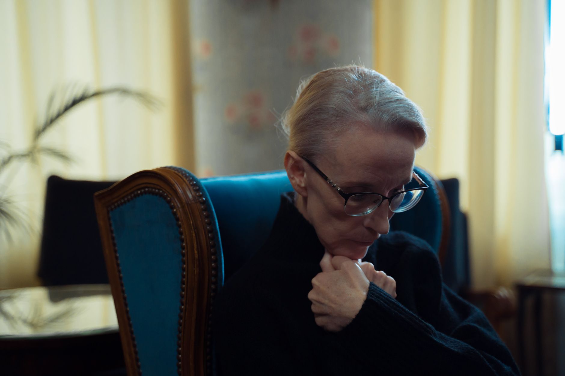 An elderly woman wearing glasses sitting in an armchair, lost in thought in a warmly lit room.