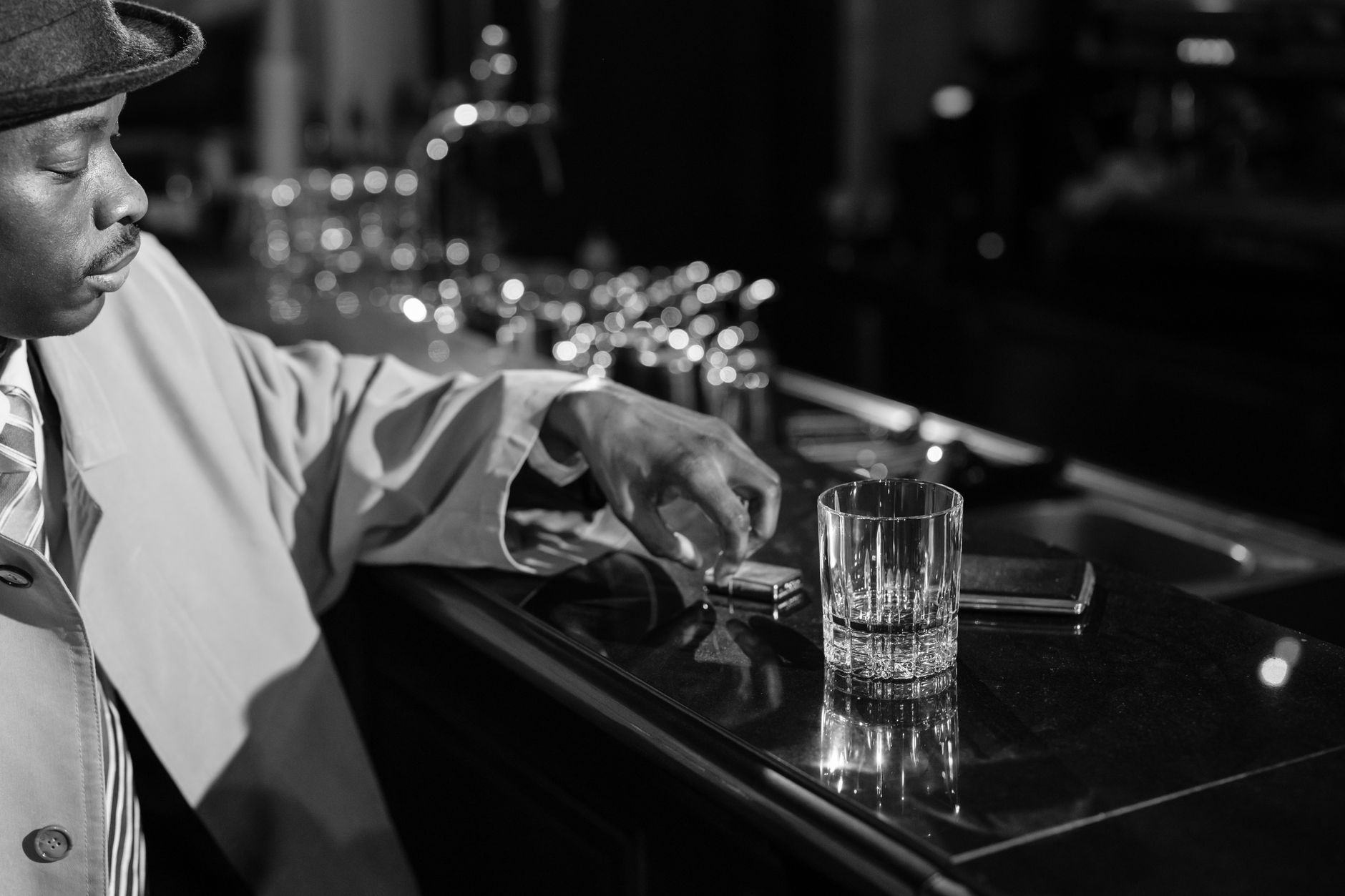 Black and white image of a man in detective attire at a dark bar counter, evoking a classic noir vibe.