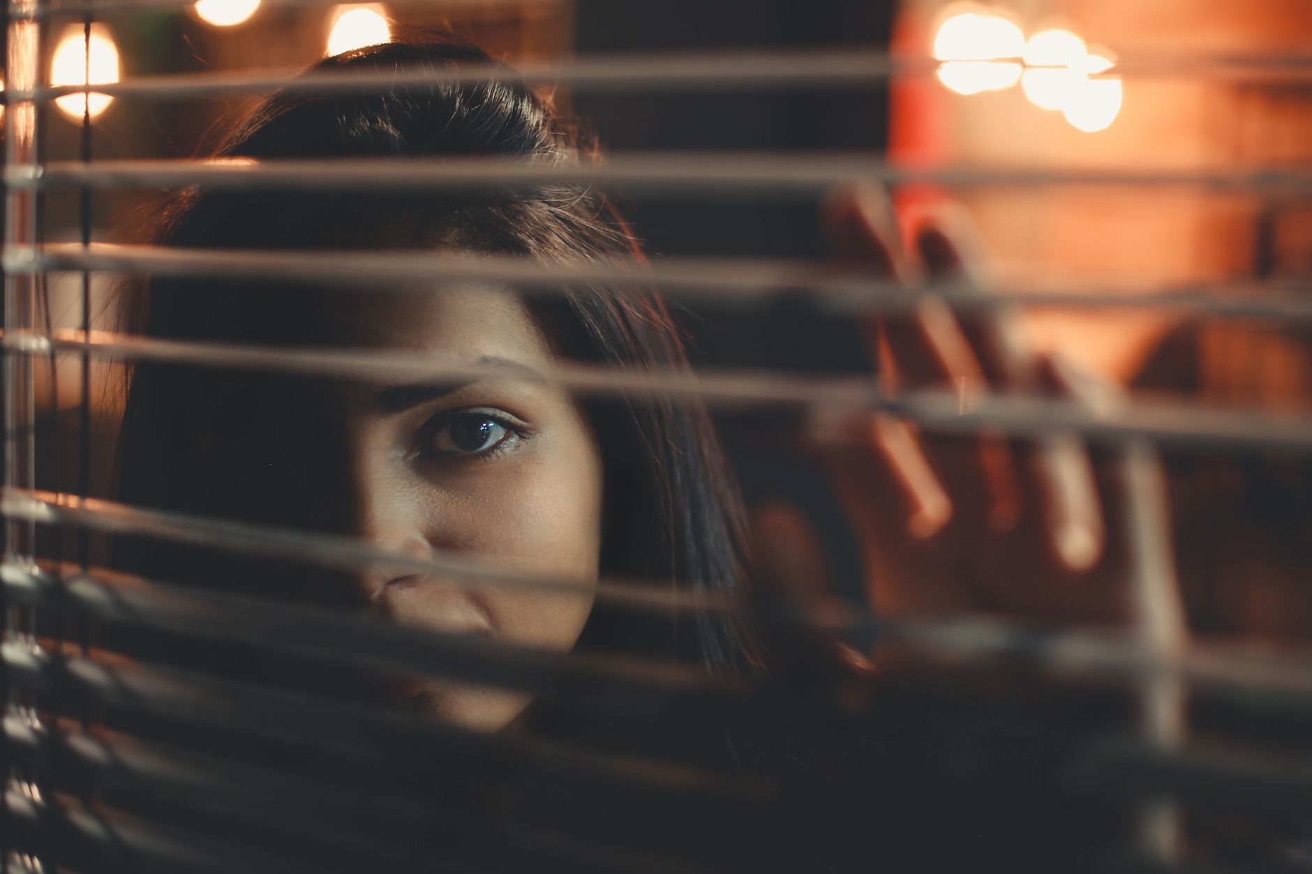 A woman peering through window blinds with a contemplative expression at night.
