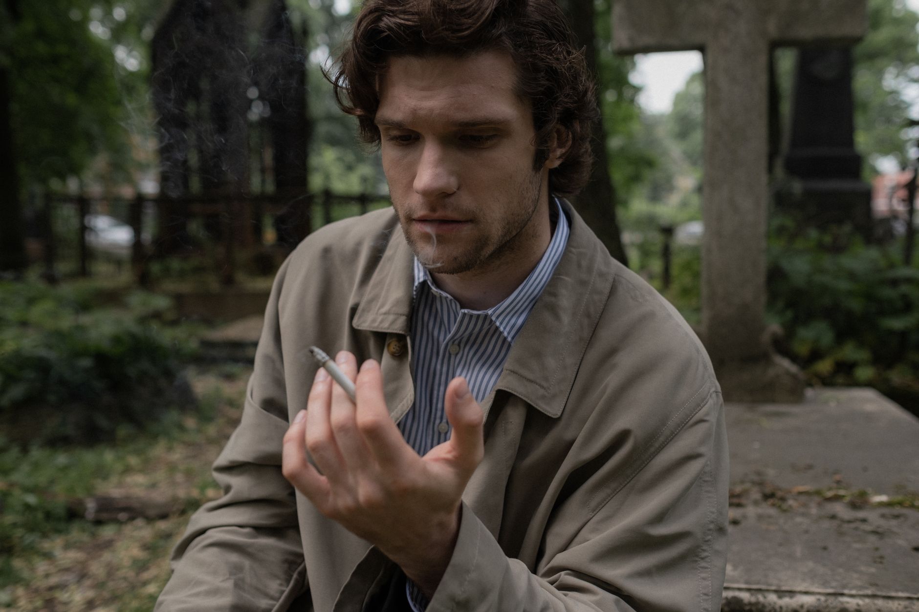 A thoughtful man in a trench coat smokes in a cemetery, evoking themes of grief and reflection.