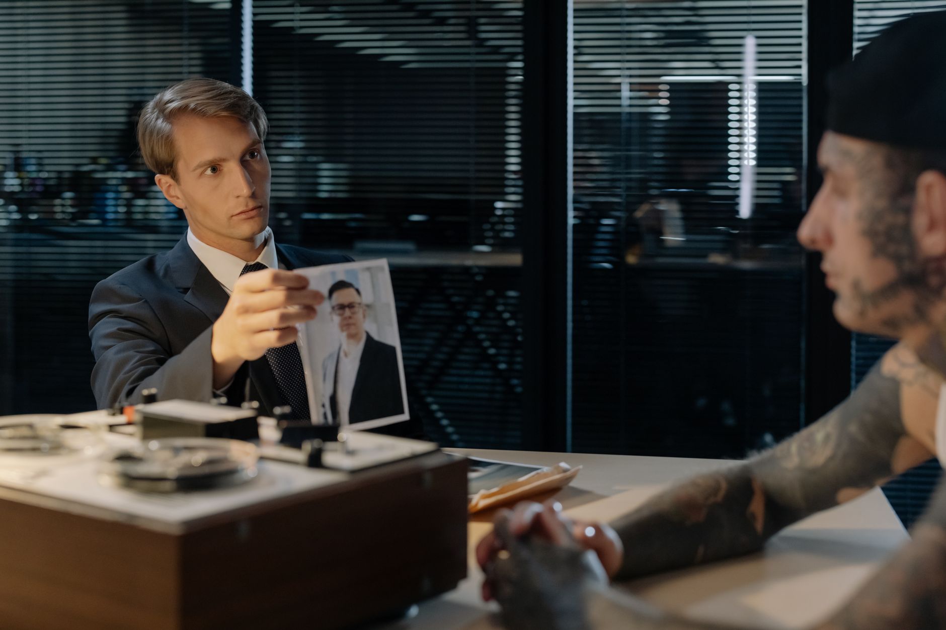 A detective interrogates a tattooed suspect in an office, examining a photograph.