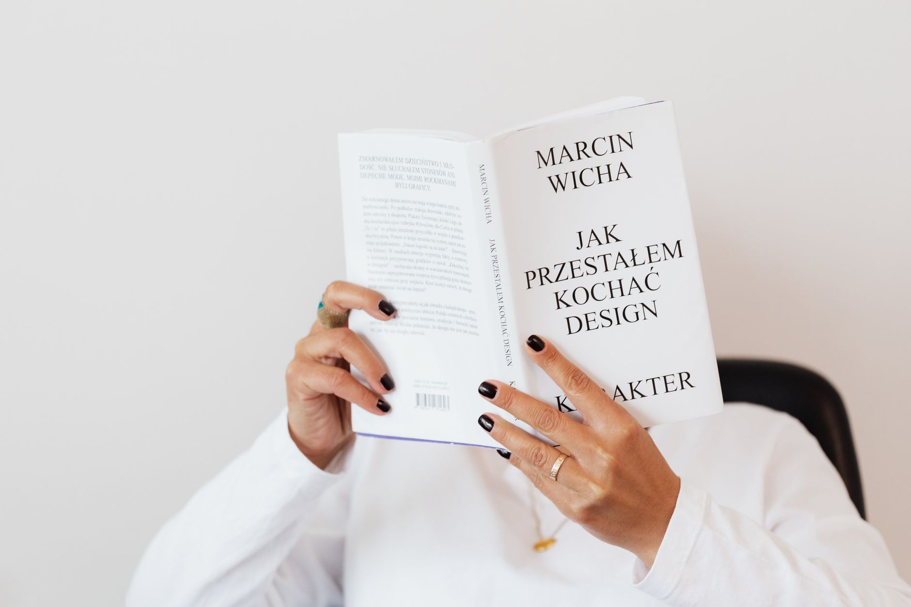 Anonymous female in white shirt with gold chain on neck reading interesting book covering face while sitting on black chair