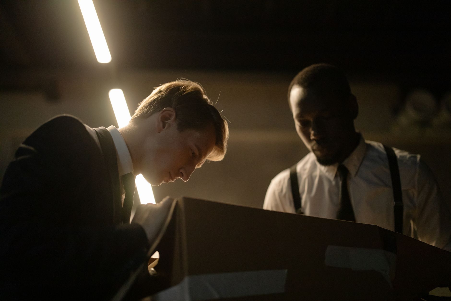 Two detectives examine a box in dimly lit room, focused on investigation.