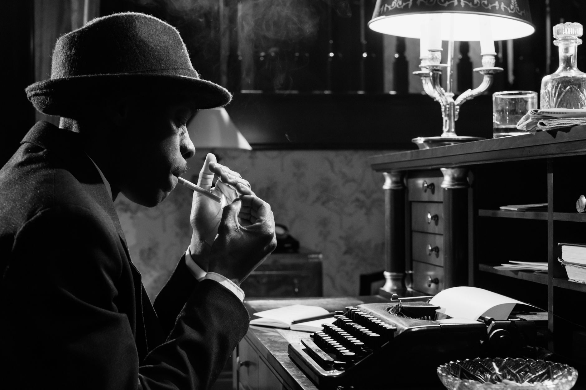 Black and white photo of a noir detective with typewriter, smoking in a vintage office setting.