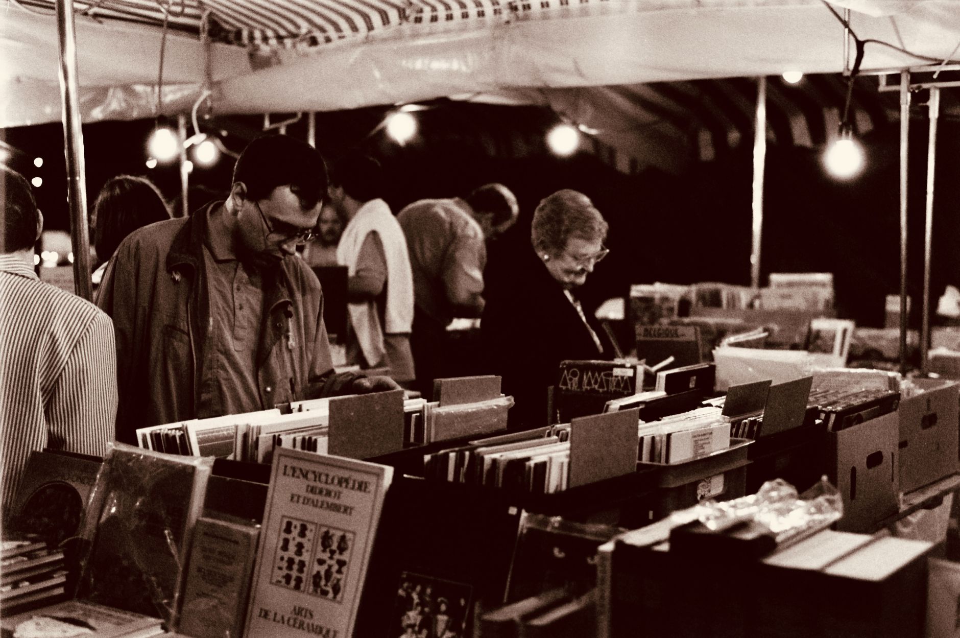 A nighttime street market scene with people browsing through book stalls in a vintage sepia tone.