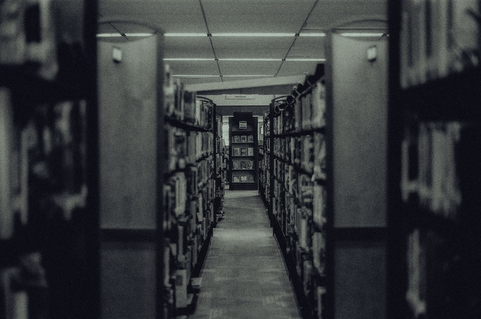 Atmospheric black and white photo capturing a library aisle with rows of shelves filled with books and a vanishing perspective.