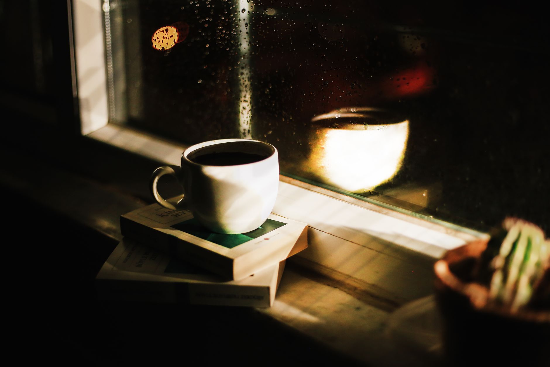 Warm coffee cup on books by a rain-streaked window, reflecting a serene indoor ambiance.