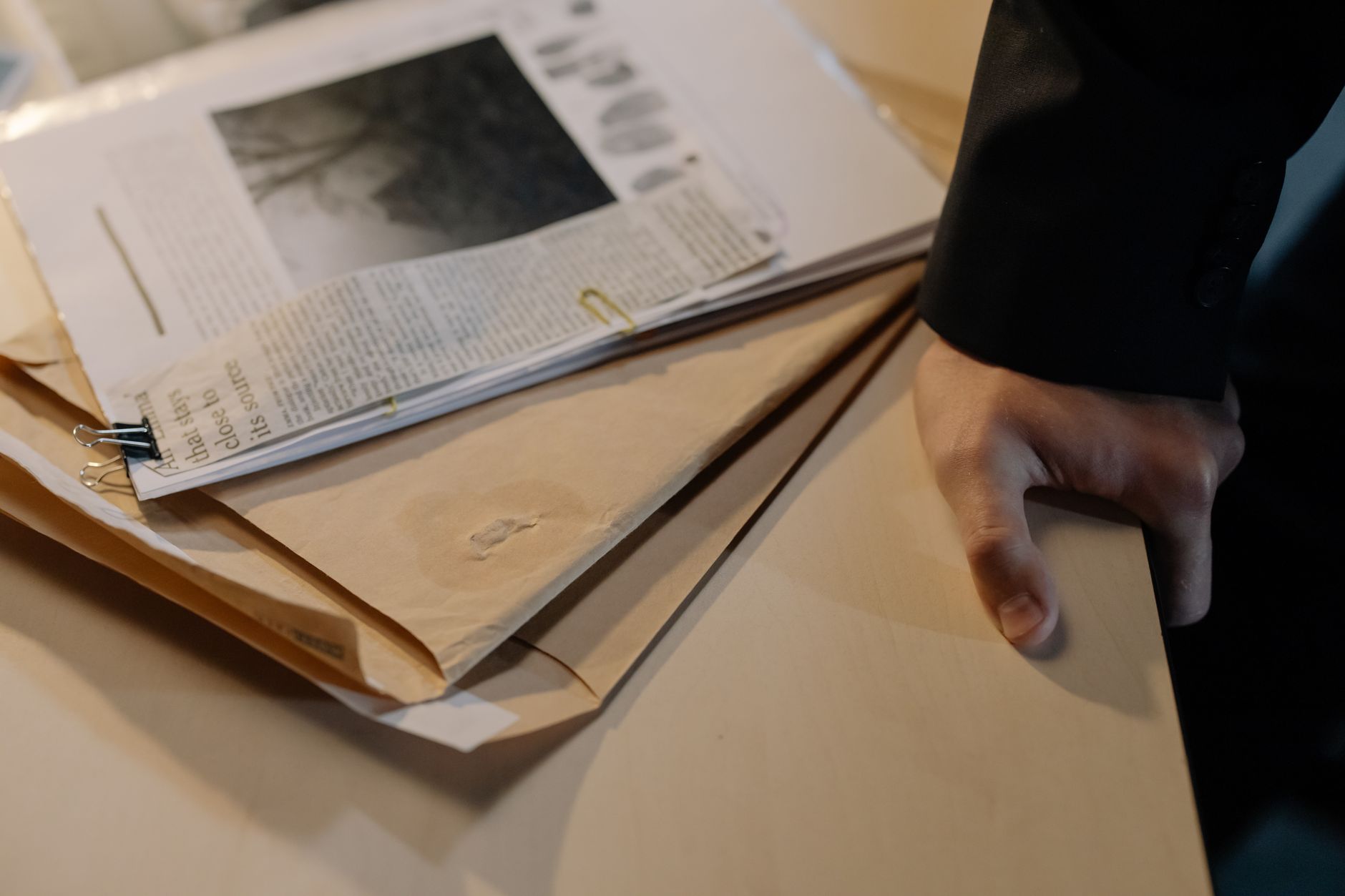 Close-up of a hand resting on a desk near a stack of investigative documents and newspaper clippings.