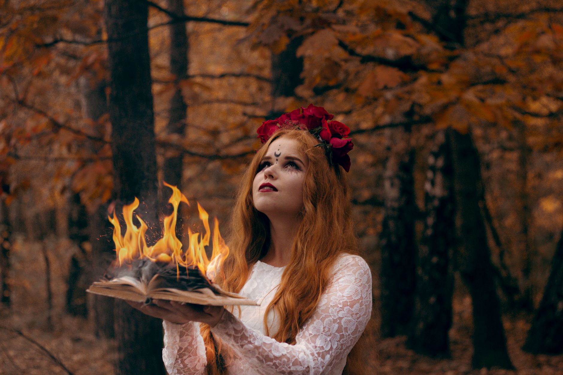 Mystical woman with red hair and flowers holds a burning book in an autumn forest.